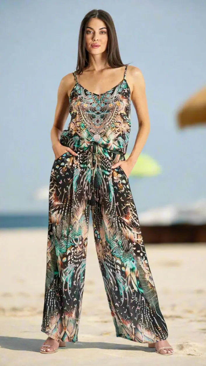 A woman stands barefoot on a sandy beach, confidently wearing the La Moda Clothing Resort Lifestyle Pant Set Cord Set featuring intricate green, black, brown, and beige patterns. She has long, straight brown hair and gazes ahead.