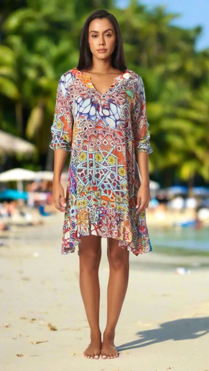 A woman stands barefoot on a sandy beach, wearing the colorful Resort Mini Dress for Beach & Beyond by La Moda Clothings. She has straight dark hair and a neutral expression, with palm trees and blurred beachgoers in the background.