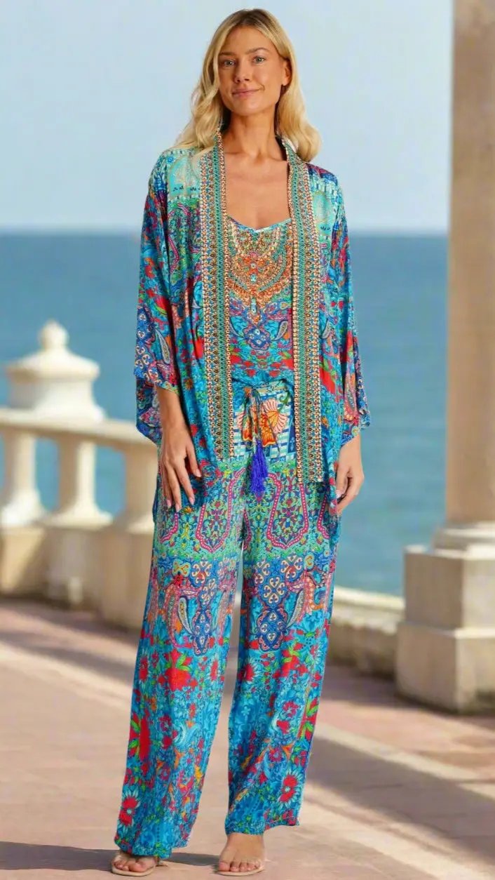 A woman with shoulder-length blonde hair smiles gently as she stands barefoot on a seaside promenade, wearing La Moda Clothing’s Resort Pant Set—an outfit featuring vibrant blue, red, and orange floral and geometric patterns.