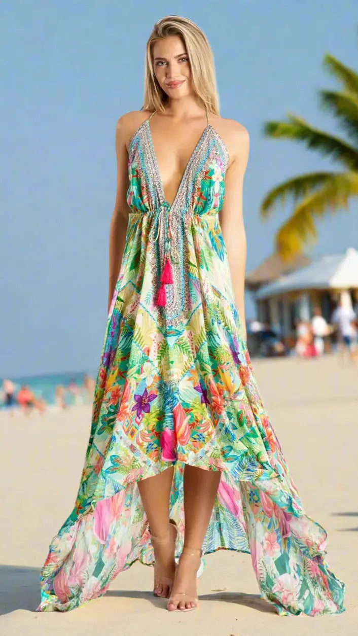 A woman with blonde hair models the La Moda Clothings Resort Wear Maxi Dress for Summer Vacations, a colorful, tropical-print design with a deep V-neck and high-low hem, standing barefoot on a beach with palm trees under a clear blue sky.