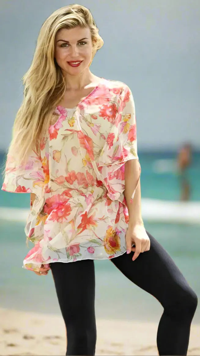 A woman with long blonde hair stands on the beach, smiling in a Lamodaclothing Sheer Chiffon Tunic—floral-patterned in pink and orange—paired with black leggings. The ocean and blurred figures are visible in the background. Look elegant this summer!.