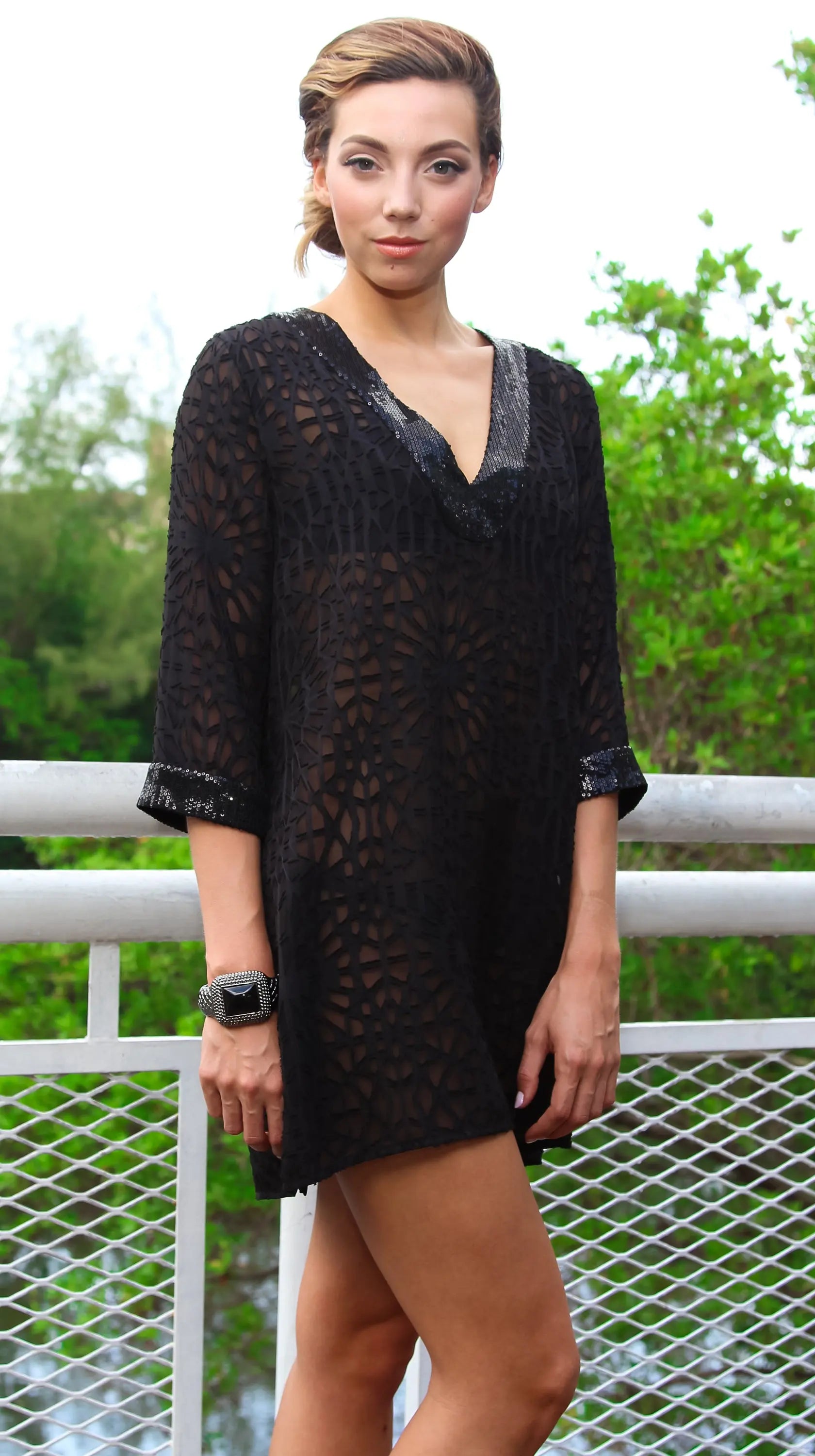 A woman with styled hair and a silver bracelet stands outdoors by a white railing, wearing the La Moda Clothing Sheer Tunic Cover Up. The black, patterned tunic features a V-neckline and three-quarter sleeves. Lush green trees fill the background.