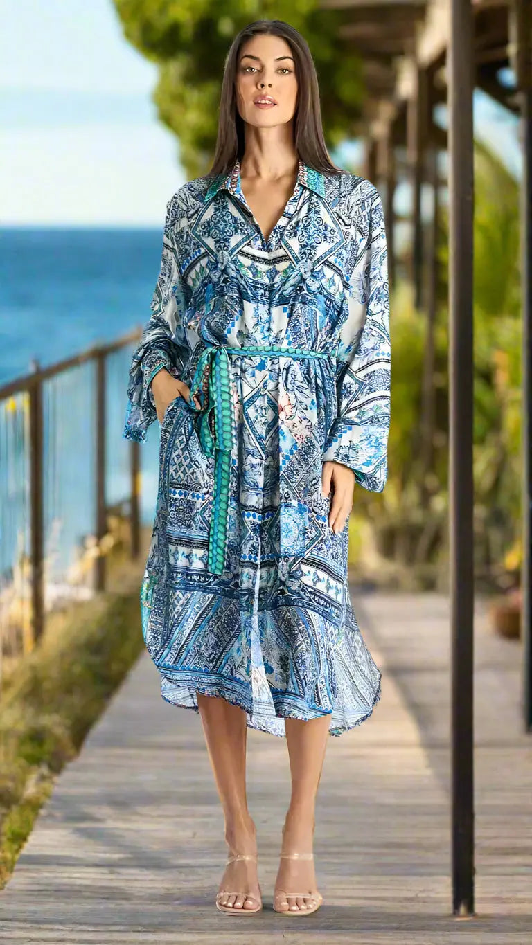 A woman stands confidently on a wooden boardwalk by the ocean, wearing La Moda Clothing’s Shop button-down shirt dress styles with clear-heeled sandals. Greenery and water form the backdrop as she gazes at the camera.