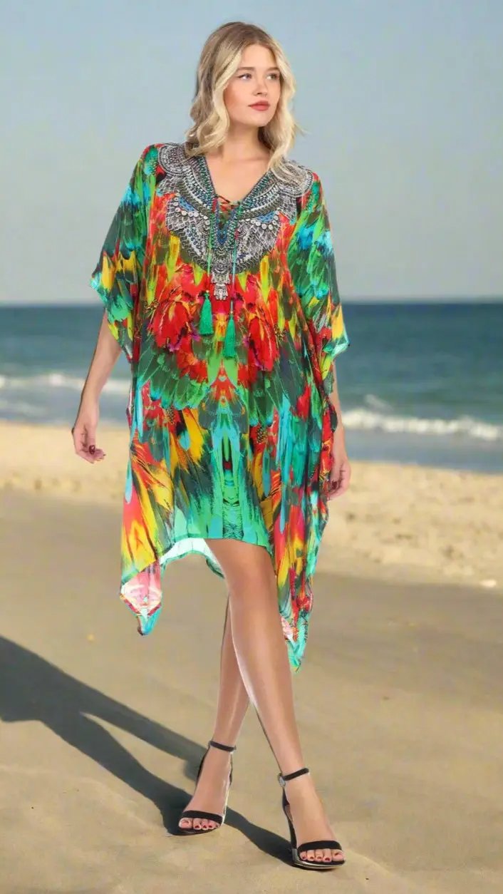 A blonde woman strolls barefoot on a sandy beach, wearing La Moda Clothings' Short Caftan Dress for Women for Summer Fun with bright red, green, yellow, and blue patterns. The ocean and blue sky are visible in the background.