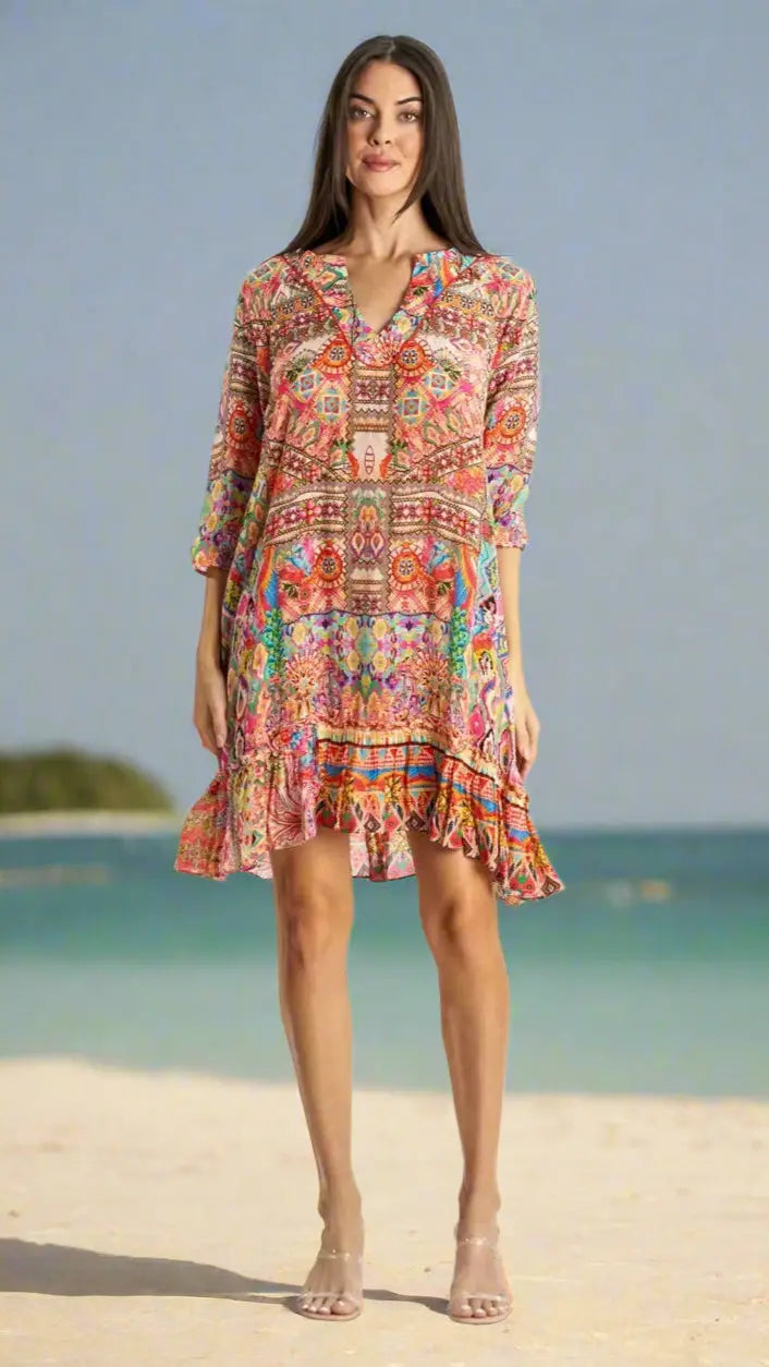 A woman stands on a sandy beach wearing the Short Dress by La Moda Clothing, featuring a colorful pattern, ruffled hem, and three-quarter sleeves. The ocean and island backdrop highlight this versatile summer style.