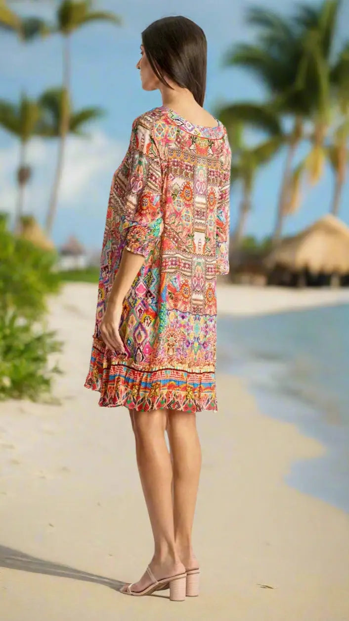 A woman in the La Moda Clothing Short Dress stands on a sandy beach with palm trees and thatched huts in the background, gazing out to sea.