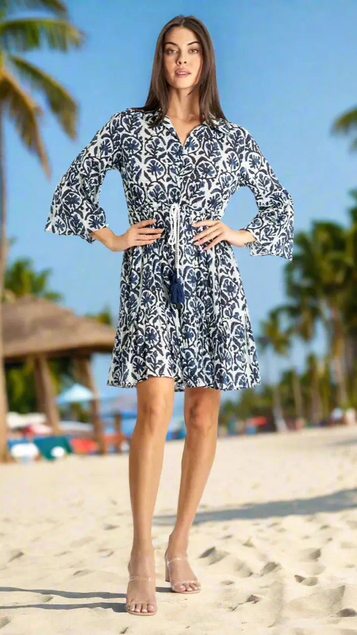 A woman with long, straight hair models the La Moda Clothing Short Dresses for Tropical Island Vacation on a sunny beach, accented by palm trees and a thatched hut in the background. She pairs the dress with clear high-heeled sandals.