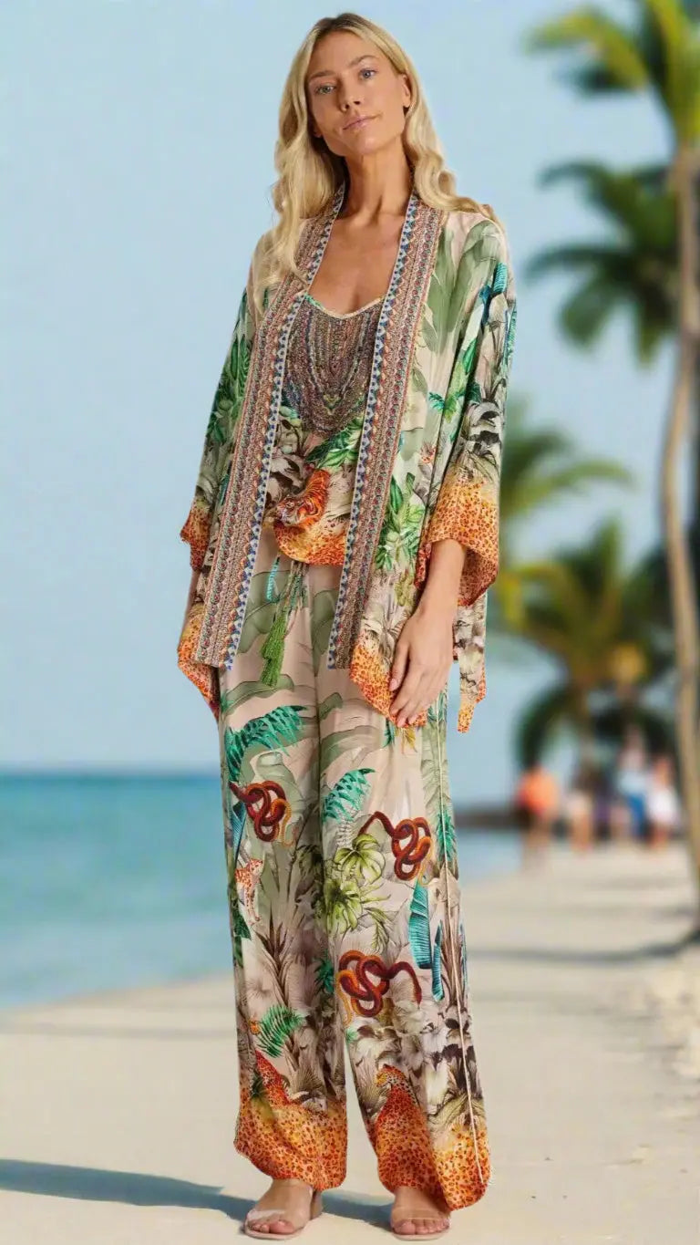 A woman with long blonde hair stands barefoot on a tropical beach, wearing La Moda Clothing's Short Kimono Cape with wide-leg pants in a bold jungle and animal print. Palm trees and blurred beachgoers are visible in the background.