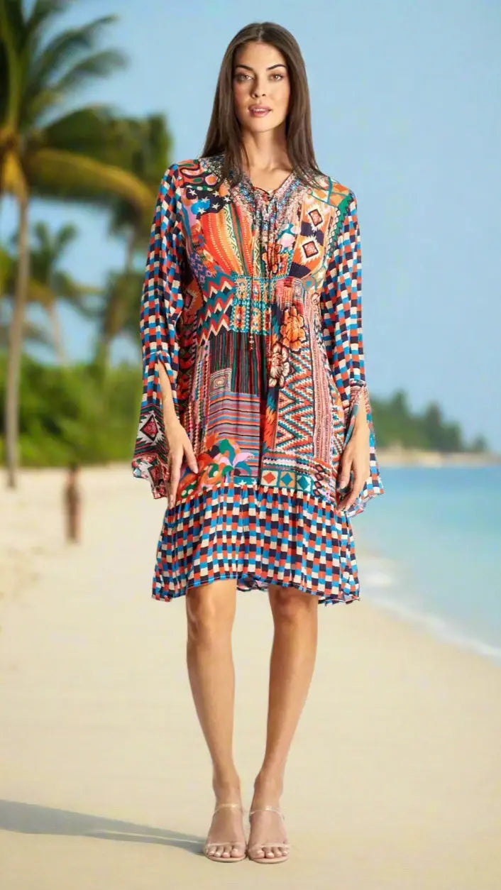A woman stands barefoot on a sandy beach with palm trees and blue water, wearing a La Moda Clothing Short Mini Dress for Summer in vibrant red, blue, orange, and white geometric and floral patterns with long flared sleeves.