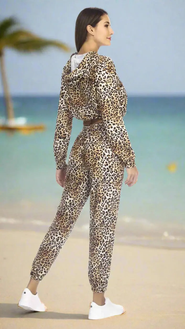 A woman with long brown hair and white sneakers stands on a sandy beach by turquoise water, wearing Club Moda Loungewear's Sporty Tie-Waist Pocket Joggers from La Moda Clothings. A palm tree and yellow kayak are in the background.
