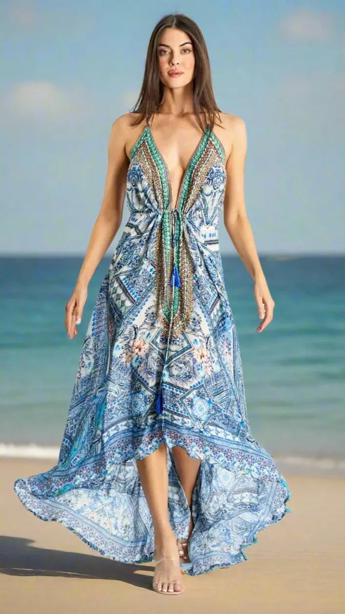 A woman with long brown hair models a flowing blue patterned halter dress from La Moda Clothing’s Summer Dresses collection, walking barefoot on a sandy beach with gentle waves and a blue sky behind her.