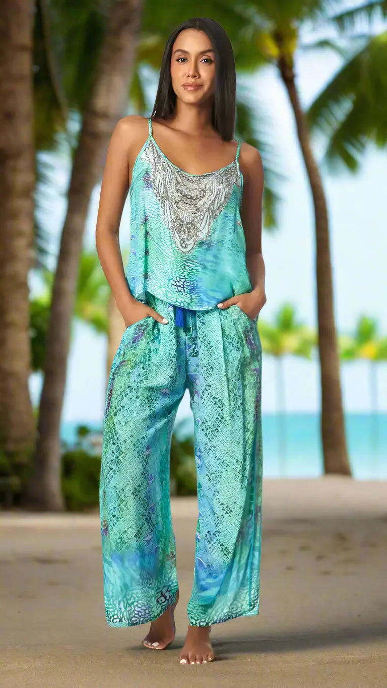 A woman stands barefoot on a sandy path lined with palms, smiling softly. She wears the La Moda Clothing Summer Fun Co Ord Pant Set in turquoise snakeskin print, with her hands in her pockets. A tropical beach and blue sky appear in the background.