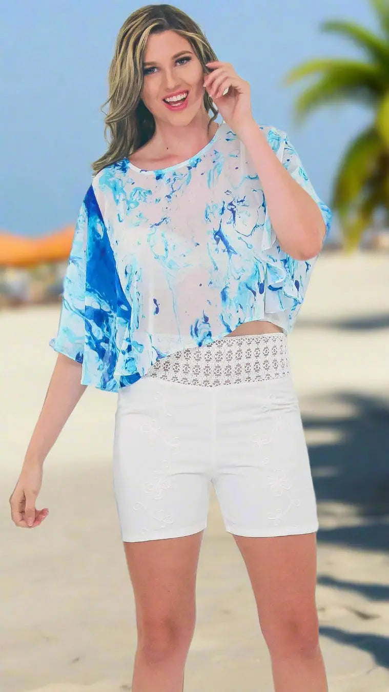 On a sunny beach, a woman smiles in Lamodaclothing’s Summer Tops in Ocean Prints, perfect for vacation, paired with high-waisted white lace-detailed shorts. Palm trees and blurred shoreline form the backdrop.