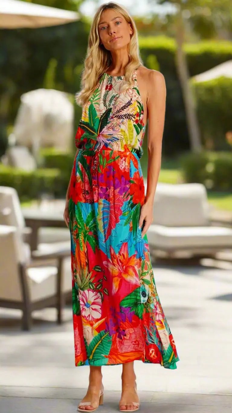 A woman with long blonde hair wears La Moda Clothing's Summertime Maxi Dress, a colorful sleeveless dress with bold floral and leaf prints, paired with nude heeled sandals. She stands outdoors with greenery and furniture in the blurred background.