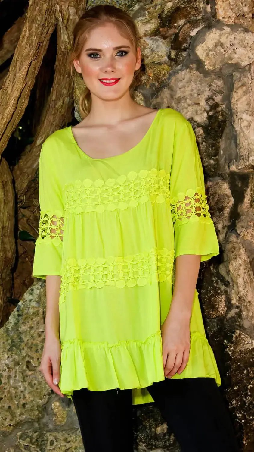 A woman with fair skin and blond hair in a ponytail stands outdoors by rocks, smiling at the camera. She wears the lamodaclothings Swimsuit Cover Up Blouse in bright yellow-green with ruffled tiers and crochet lace, plus black leggings.