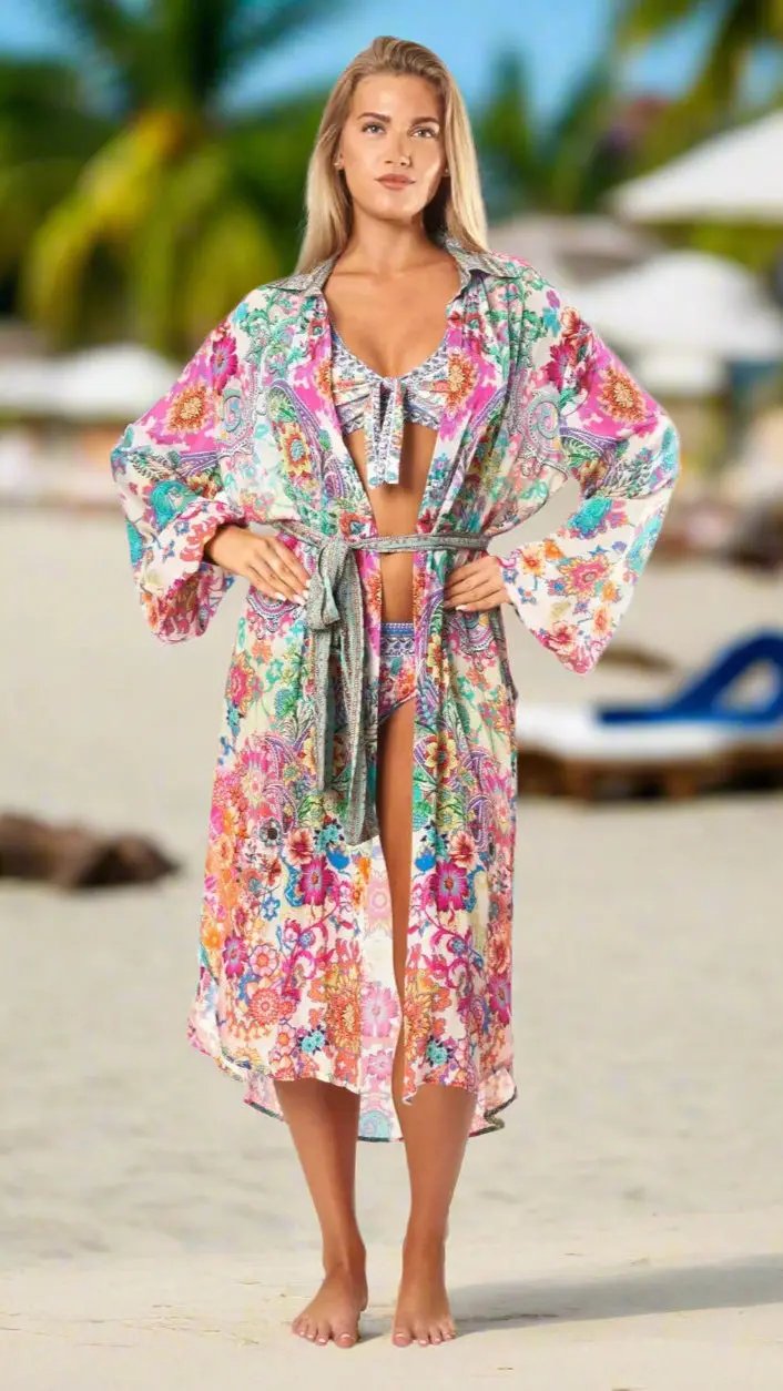 A woman stands barefoot on a sandy beach, confidently wearing The Effortless Shirtdress Cover-Up by La Moda Clothings over her bikini. Palm trees and lounge chairs blur in the background as sunlight brightens her face and hair.