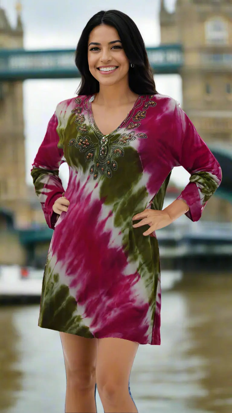 A woman with long dark hair smiles outdoors in front of London's Tower Bridge, wearing La Moda Clothing's Tie Dye Knit Tunic in maroon and olive green, featuring long sleeves and beaded neckline detailing.