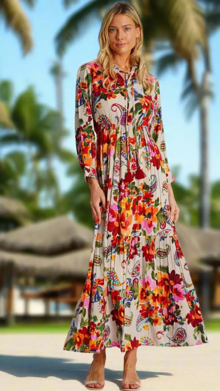 A woman with blonde hair smiles barefoot on the sand in a long, colorful La Moda Clothing Trending Bohemian Maxi Dress. Palm trees and thatched-roof huts in the background suggest a tropical vibe.