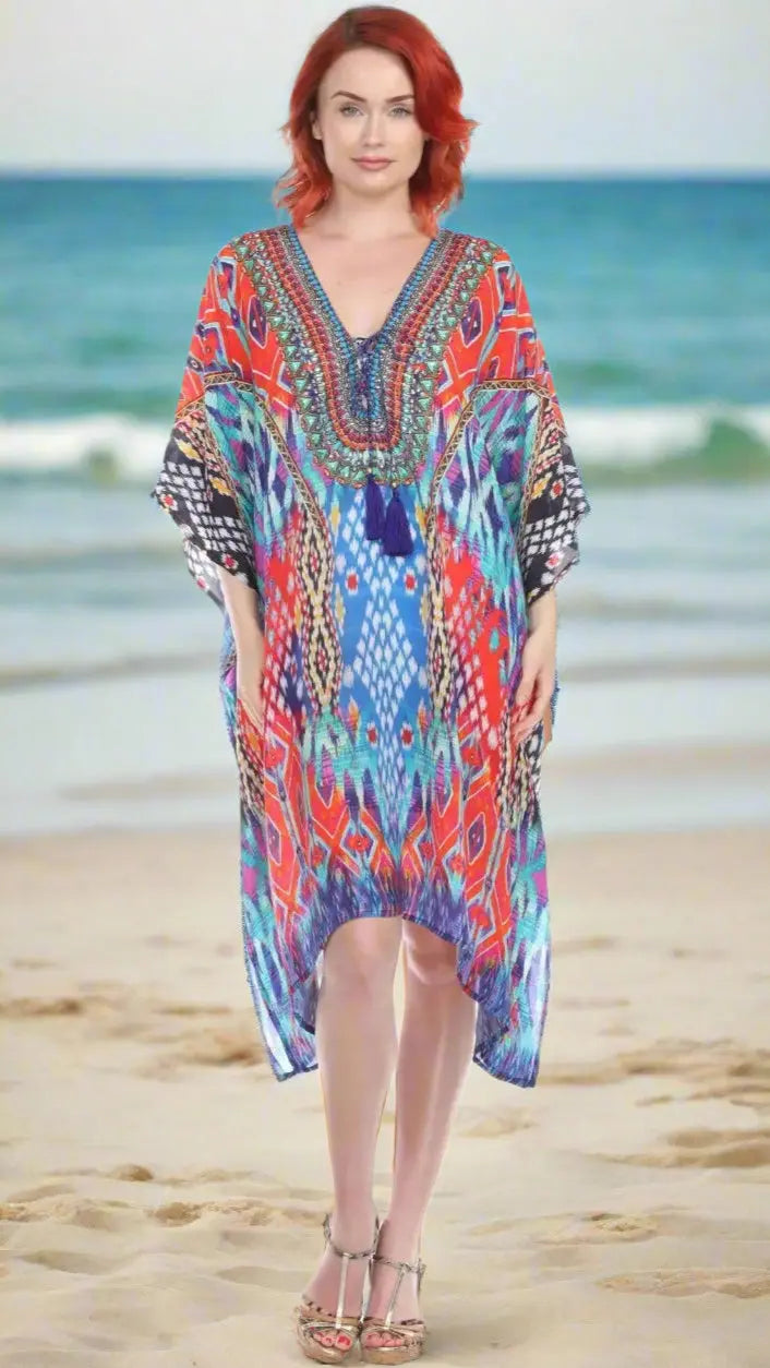 A red-haired woman stands barefoot on the sand by the ocean, smiling in a colorful Trendy V-Neckline Luxury Kaftan Dress with blue and red geometric patterns from Lamodaclothing.