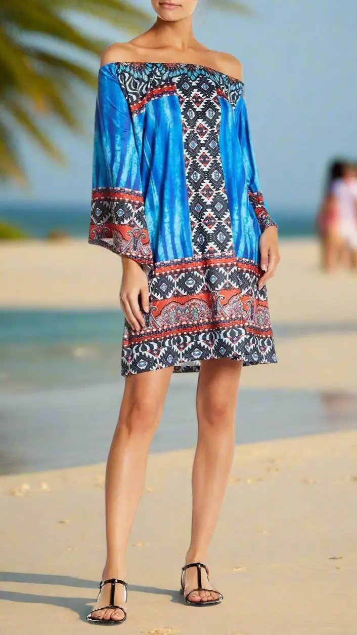 A woman stands barefoot on a sandy beach in the lamodaclothings Tunic Style Off-Shoulder Beach Dress with blue and red patterns. Palm leaves and two blurred figures appear in the background under a bright sky.
