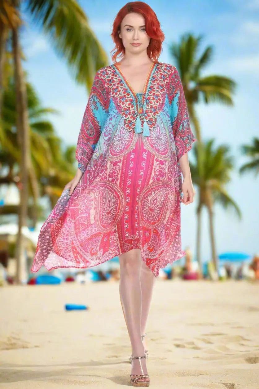 A red-haired woman stands barefoot on a sandy beach with palm trees, smiling in a Lamodaclothing Viscose Silk Designer Kaftan Dress featuring vibrant pink, red, and blue patterns under a sunny sky.