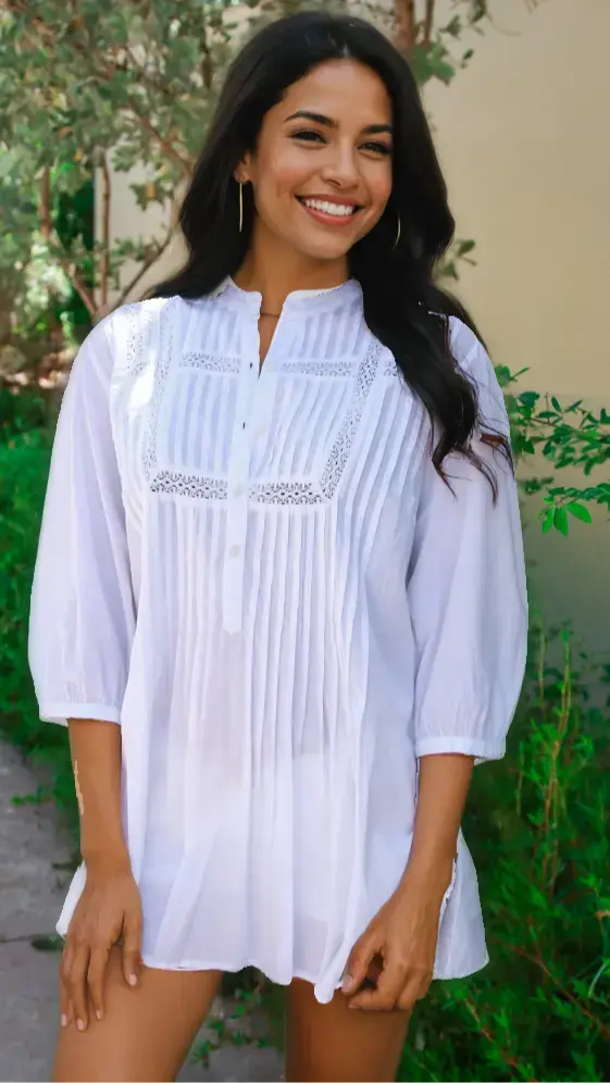 A woman with long dark hair smiles outdoors, posing in front of greenery and a light wall while wearing the La Moda Clothing White Cotton Tunic featuring lace and pleat details on the front.