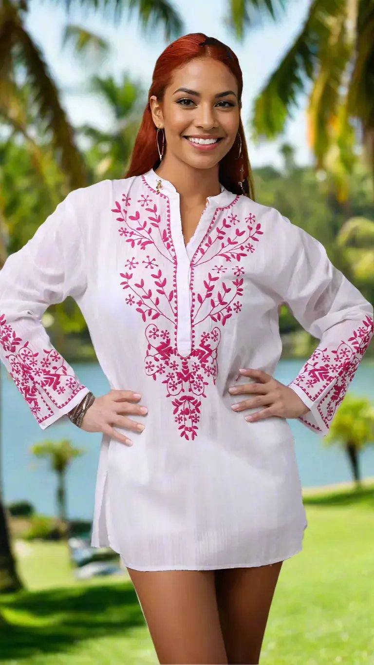 A smiling woman with long, straight red hair stands outdoors wearing La Moda Clothing’s White Embroidered Tunic. The white tunic features intricate pink embroidery on the chest, sleeves, and cuffs. Palm trees and water blur in the sunny background.