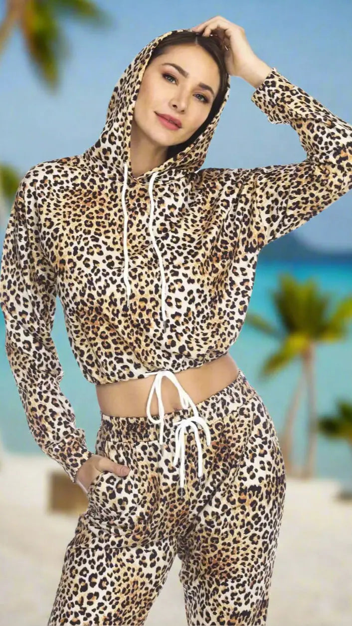 A woman poses on a tropical beach wearing the Women's Bryce Cropped Hoodie in Leopard Print by La Moda Clothings, with palm trees and turquoise water blurred in the background.