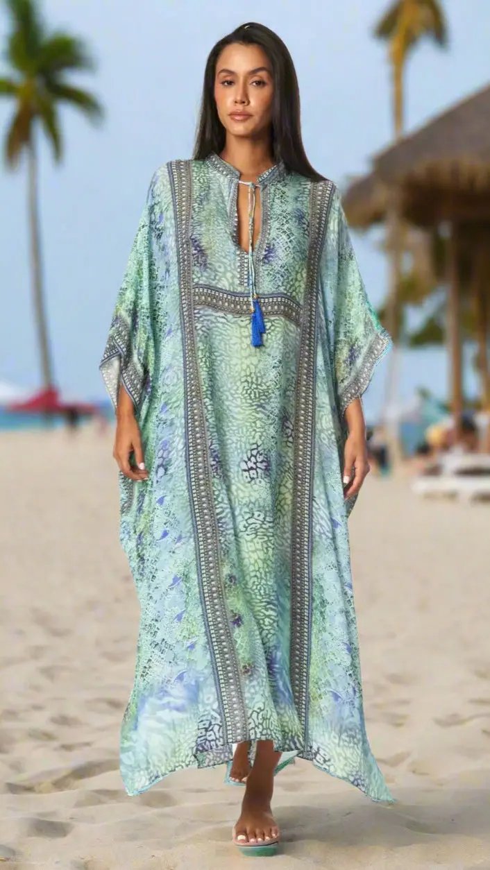 A woman stands barefoot on the sand in a La Moda Clothings Women's Designer Maxi Caftan Pool Party Gown Dress, its light green and blue patterns flowing by palm trees and a thatched hut, setting a tropical, relaxed scene.