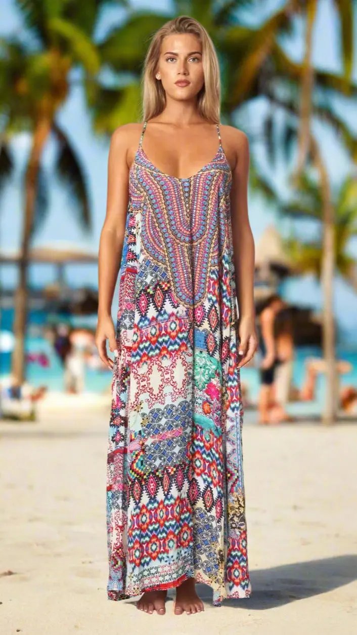 A woman with long blonde hair stands barefoot on a beach in a La Moda Clothings Women’s Maxi Dress with Pockets, featuring colorful patterns and thin straps. Palm trees and blurred people appear in the sunny, tropical background.