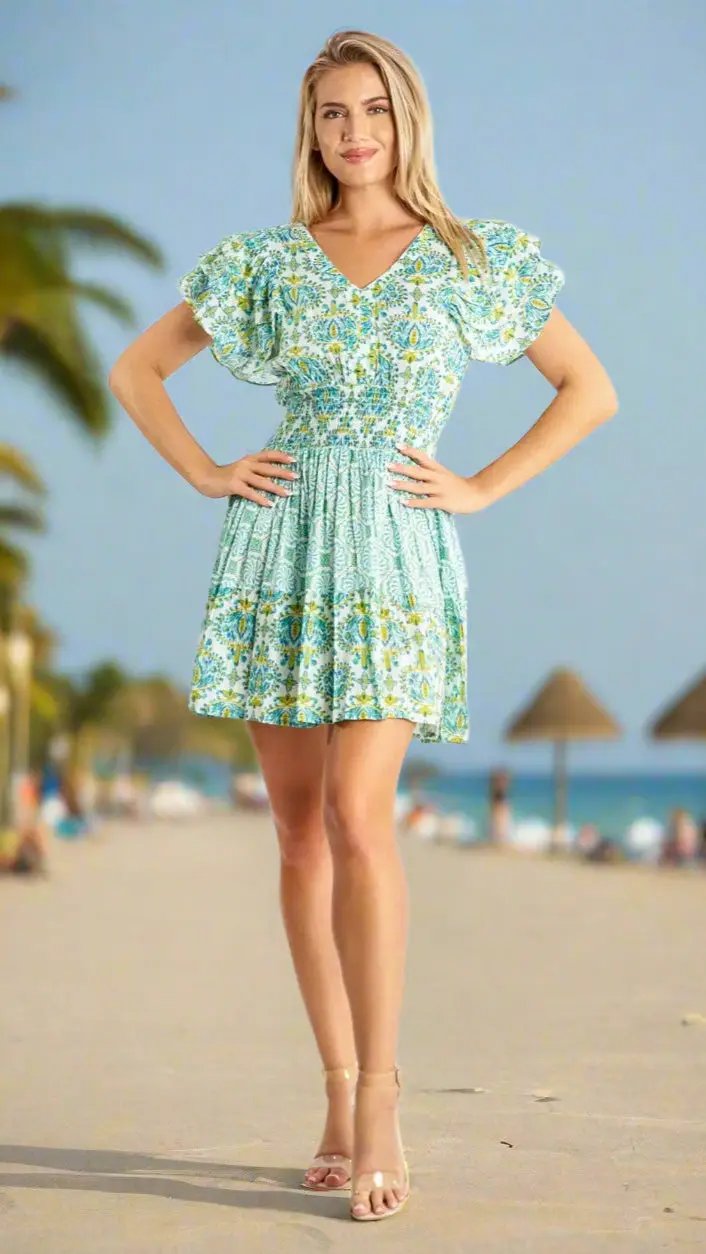 A woman stands on a sunny beach wearing the La Moda Clothing Mini Beach Dress, a short light blue floral dress with ruffled sleeves. She has long blonde hair, nude sandals, and poses with hands on hips; palm trees and umbrellas behind her.